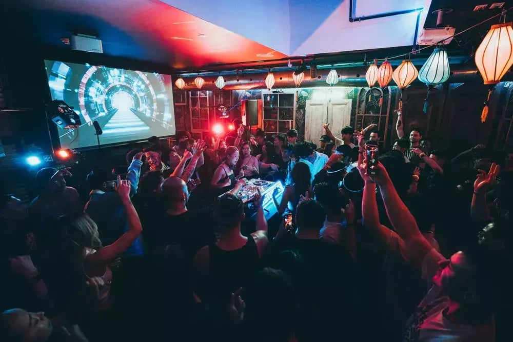 A female DJ is seen performing in a club or bar with a screen behind her, while people celebrate and record her with their smartphones.