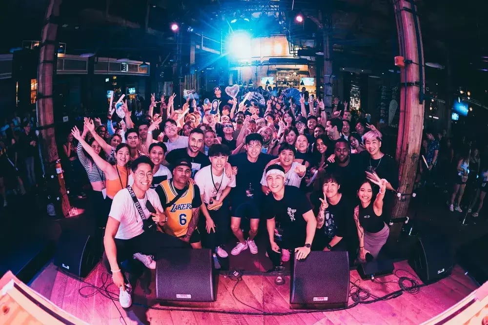 A group of people on stage pointing towards a screen, with a crowd smiling and posing for the camera, some raising their hands. The shot takes place in a nightclub or concert hall.